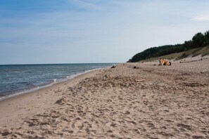 Ein sandiger Strand mit ruhiger See, einige Personen und ein bewaldeter Hügel im Hintergrund.