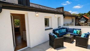 Une terrasse extérieure avec un canapé et des fauteuils, une petite table et une porte donnant sur un espace intérieur.