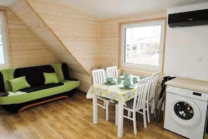 A compact room with a wooden sloped ceiling, a green sofa with cushions, a dining table with chairs, and a washing machine. - Apartment in Wisełka with a large terrace (Wisełka)