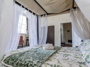 Un dormitori amb un llit, una finestra amb cortines translúcides, una porta i una taula de nit.