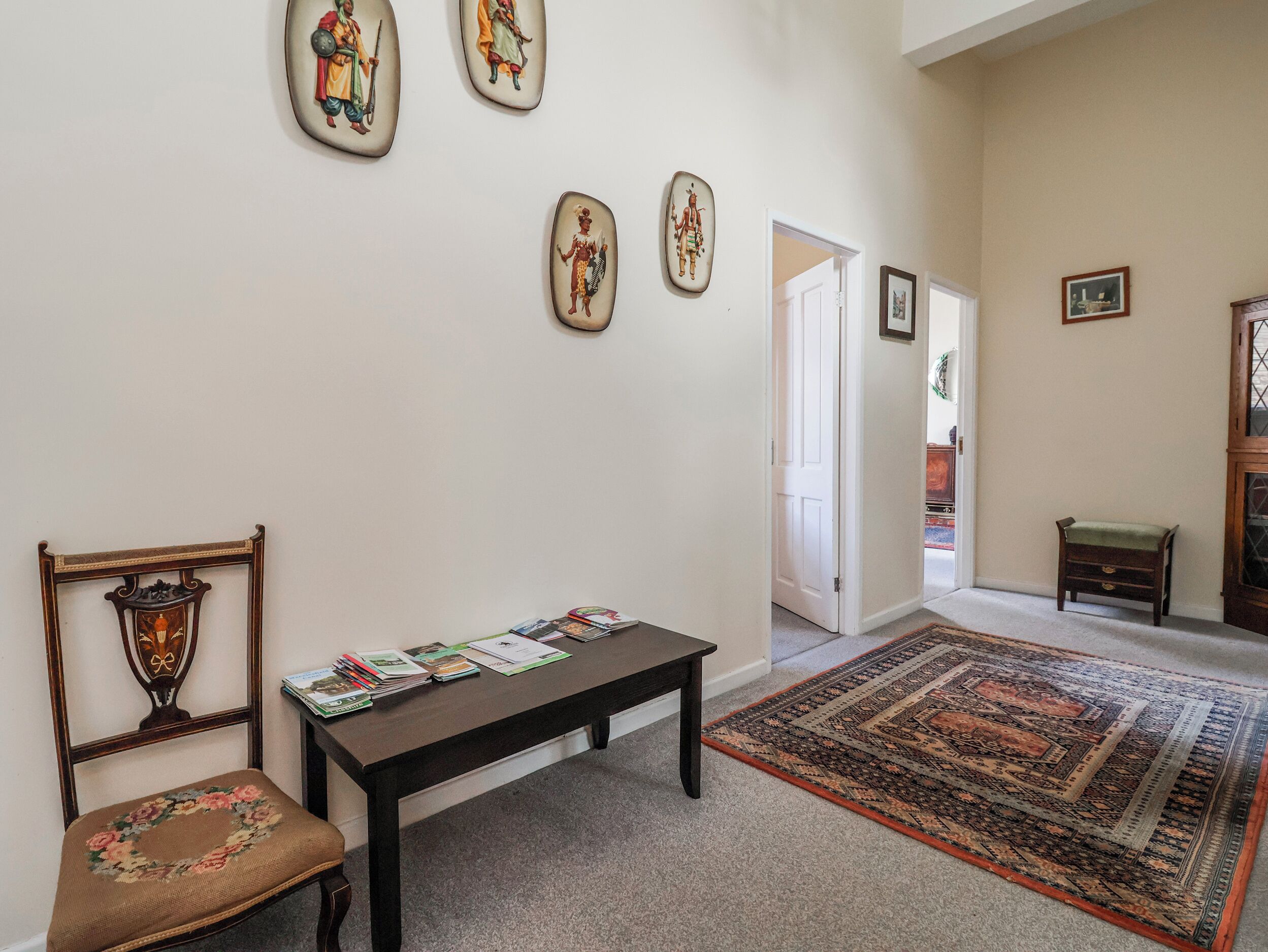 Una habitación con un banco de madera, una silla con cojín estampado, una mesa con revistas y una alfombra con motivos, además de obras de arte colgadas en la pared.
