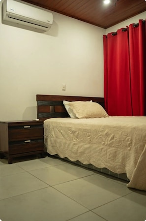 A bedroom with a bed, a nightstand, a wooden dresser, a red curtain, and an air conditioning unit.