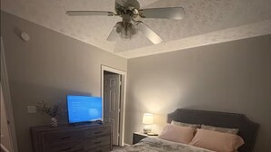 Ett sovrum med en säng, ett nattduksbord, en lampa, en tv på en byrå, en takfläkt och ett fönster.
