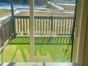 On aperçoit, à travers une porte-fenêtre coulissante, un jardin avec une terrasse en bois, une pelouse verdoyante et, au fond, une zone recouverte de neige.