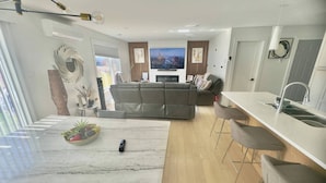 Una sala de estar moderna con un sofá, una mesa de comedor con un cuenco de fruta y una cocina con un lavaplatos y taburetes de bar.