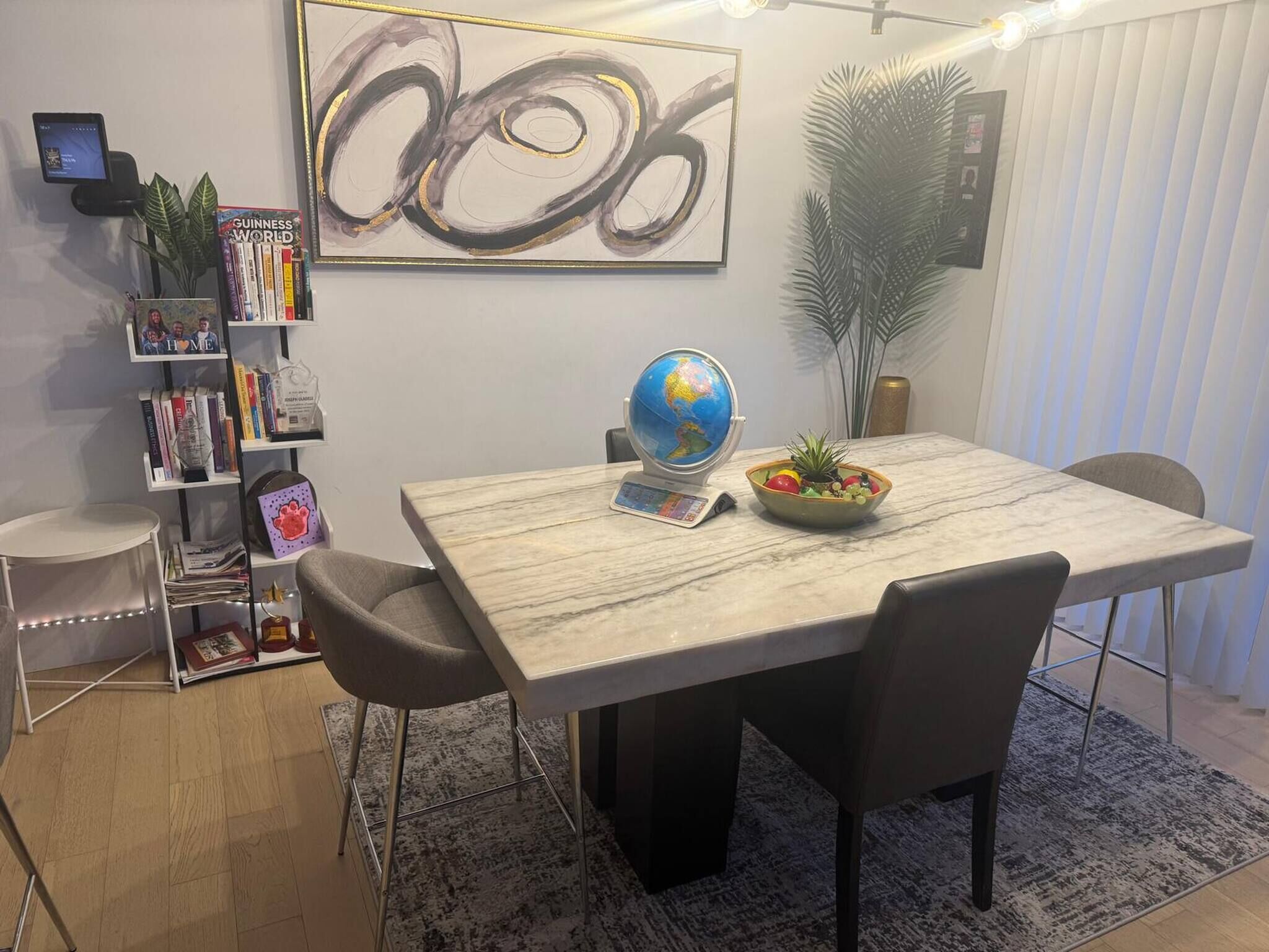 Un comedor con una mesa de mármol, sillas, un globo terráqueo, un cuenco de fruta, una estantería y un cuadro abstracto de gran tamaño.