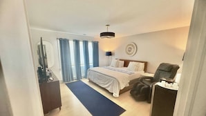 Habitación de hotel con cama, escritorio, silla, sillón de masaje, lámpara y vistas al exterior.