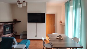 A modern living room with a fireplace, a flat-screen TV mounted on the wall, a dining table with chairs, and a large window with curtains. - Green Ledro Lake Apartment -A few steps from the Ponale Trail - 800m from the Lake (Molina di Ledro)