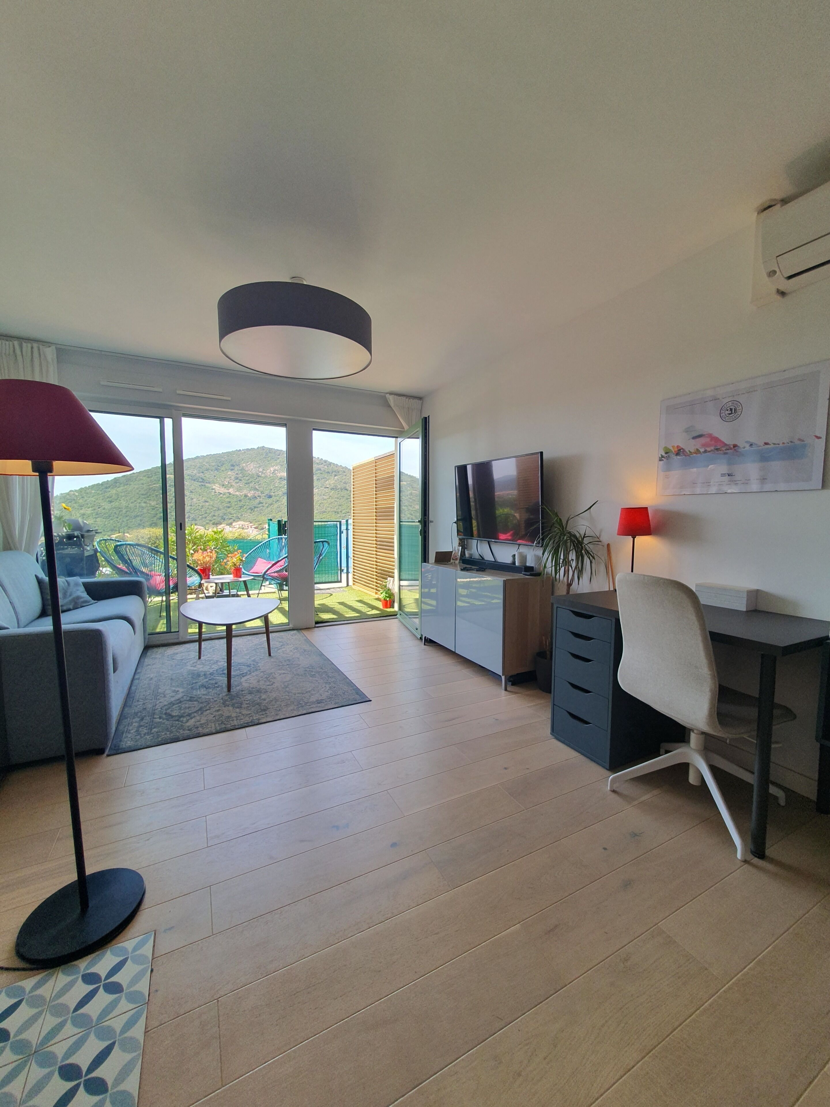 A modern living room with a sofa, a desk, a TV, and a view of a garden and hills.