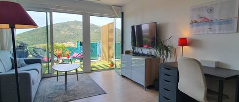 Ein modernes Wohnzimmer mit Sofa, Schreibtisch, Fernseher und Blick auf Garten und Hügel.