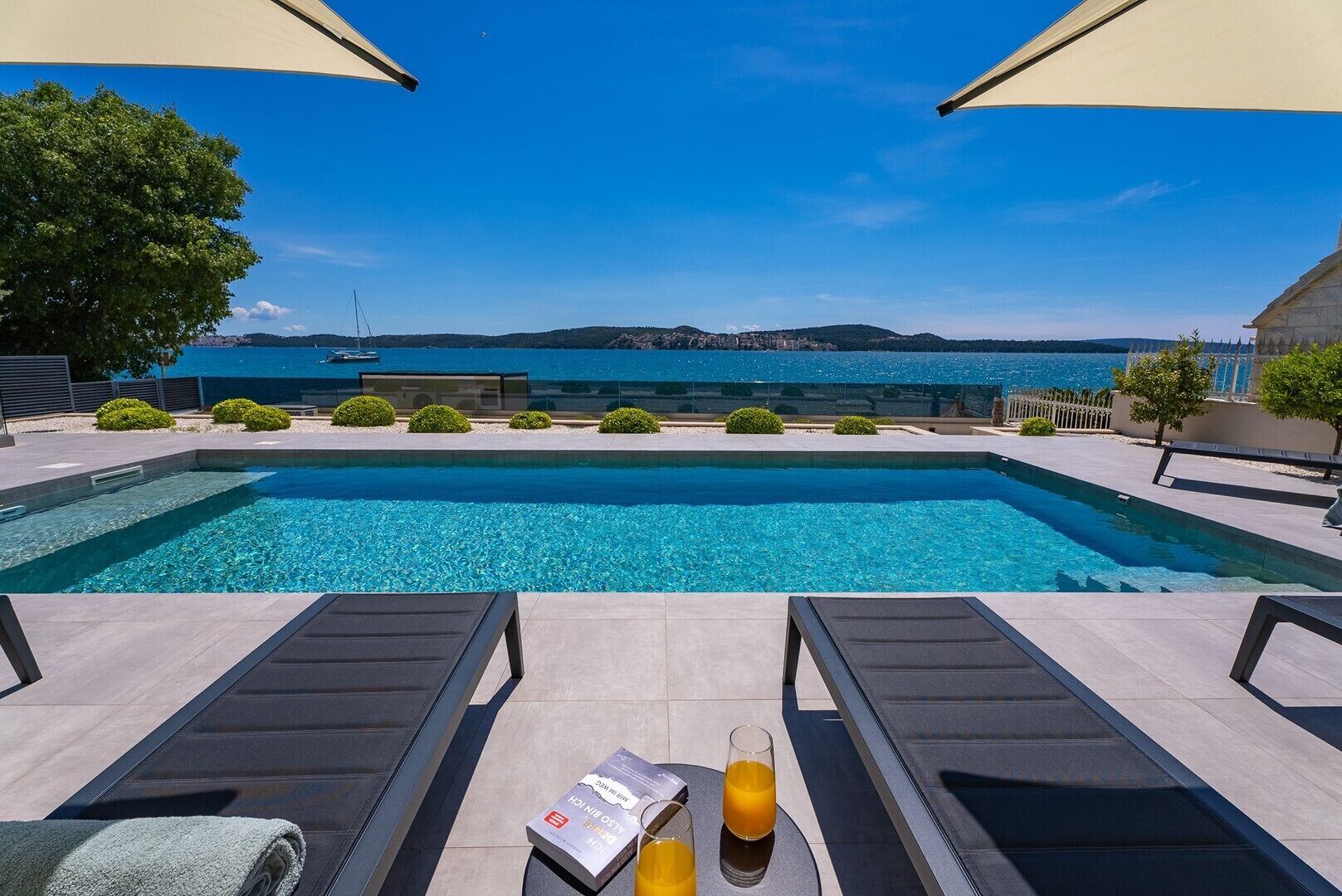 Uma piscina com água azul cristalina, cadeiras de descanso, uma mesa com toalha e um livro, e vista para o mar e colinas ao longe.