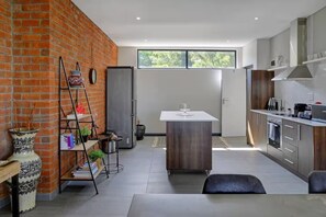 A modern kitchen with a brick wall, a central island, and a dining area with a table and chairs. - Stylish Penthouse Heart of the City by Ctha (Cape Town)