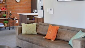 Un sofá de cuero marrón con cojines estampados, una pared de ladrillo, una mesita con una lámpara y una estantería con objetos decorativos.