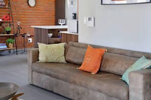 A brown leather sofa with patterned cushions, a brick wall, a small table with a lamp, and a bookshelf with decorative items. - Stylish Penthouse Heart of the City by Ctha (Cape Town)