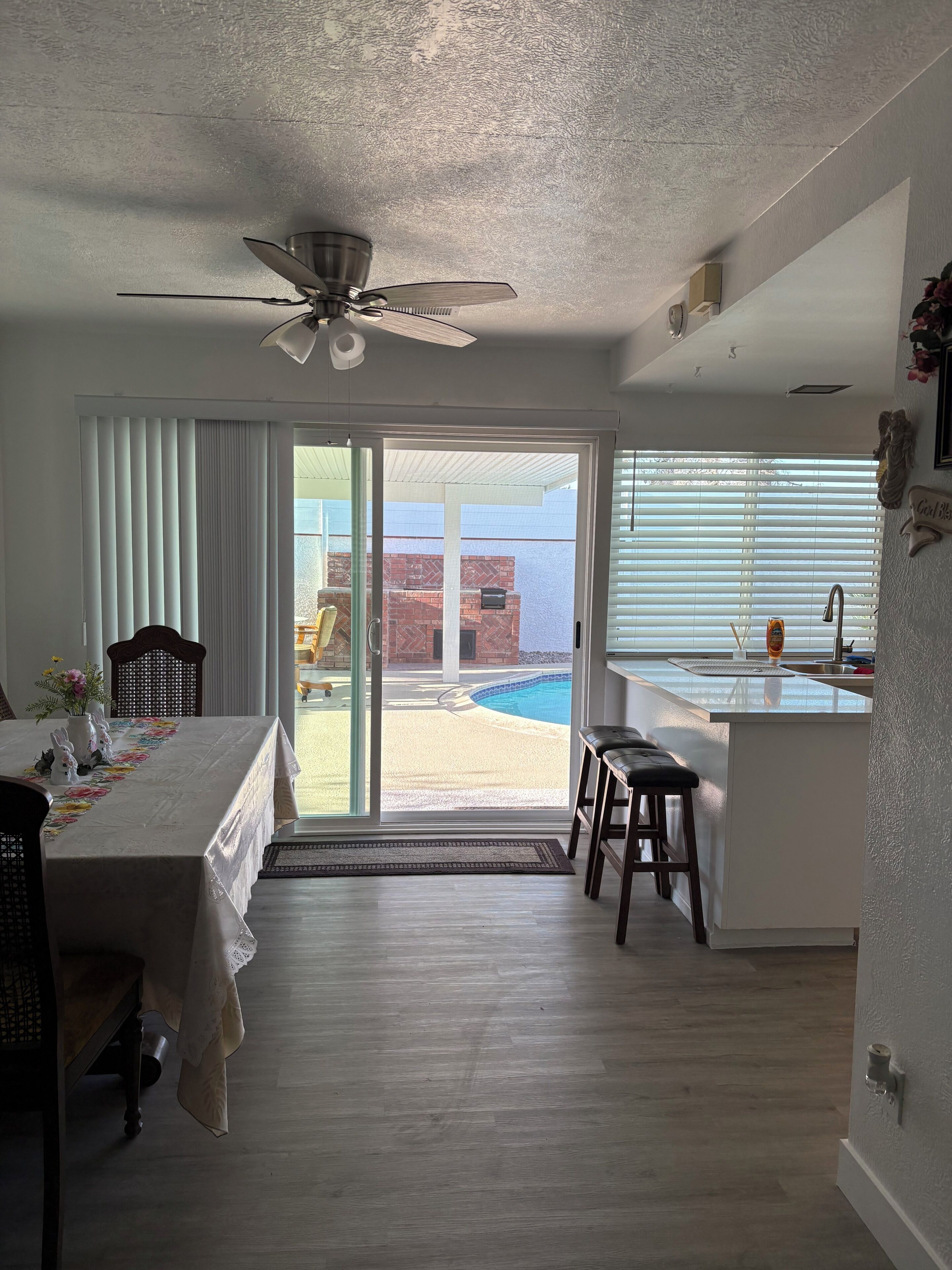 Una habitación con una mesa de comedor, sillas, un ventilador de techo, una zona de cocina y un ventanal corredizo que da a un patio exterior con piscina.