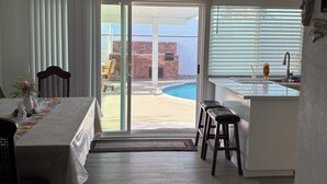A room with a dining table, chairs, a ceiling fan, a kitchen area, and a sliding glass door leading to an outdoor patio with a pool.