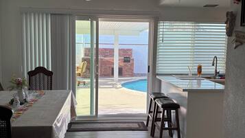 Una habitación con una mesa de comedor, sillas, un ventilador de techo, una zona de cocina y un ventanal corredizo que da a un patio exterior con piscina.