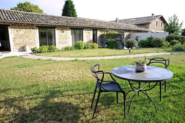 A stone building with a tiled roof, a paved pathway, and a round table with chairs in the foreground.