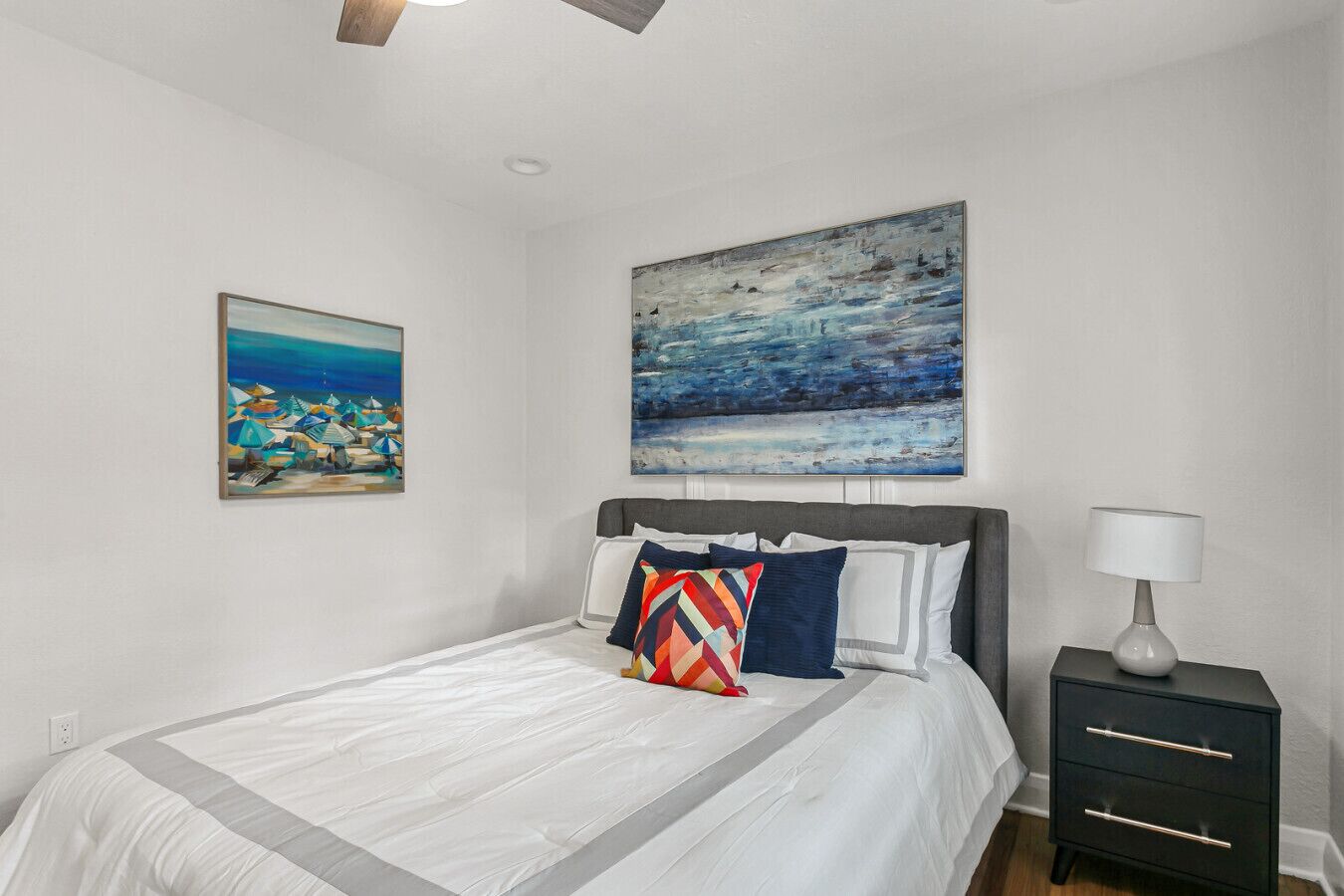 Um quarto com uma cama, uma mesinha de cabeceira, um abajur e duas pinturas na parede.