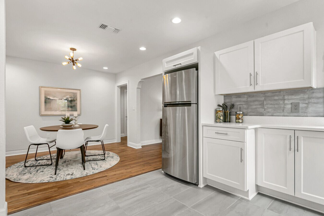 A modern kitchen with white cabinets, a stainless steel refrigerator, and a dining area with a round table and chairs.