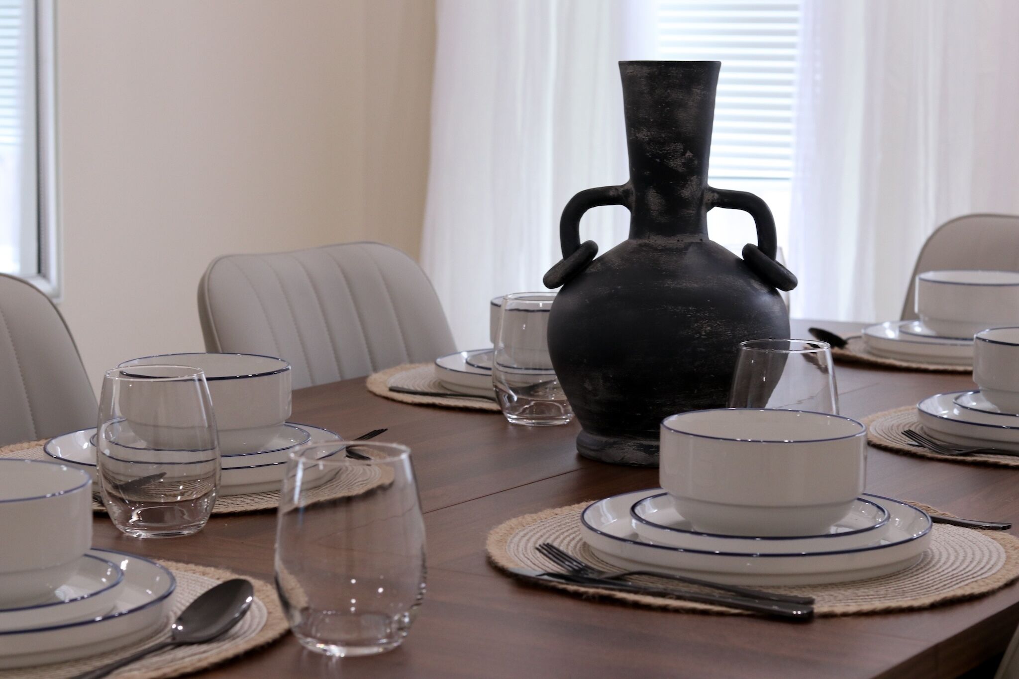 A dining table set with plates, bowls, glasses, and cutlery, with a large vase in the center.