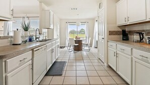 Una cocina con armarios blancos, una mesa de comedor con sillas, vista a través de la ventana y una cafetera sobre la encimera.