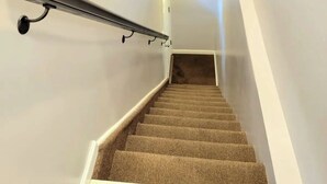 Una escalera con peldaños alfombrados, barandilla de metal y paredes blancas.
