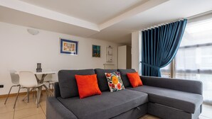 Un salon moderne avec un canapé d’angle gris, deux coussins orange, un coussin coloré, une table à manger blanche avec des chaises et une fenêtre avec des rideaux bleus.