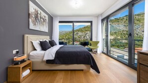 A modern bedroom with a large bed, a bedside table, a chair, and a balcony with a view of the landscape. - Luxury Five Bedroom Villa With Pool (Zaton)