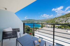 Ein Balkon mit Blick auf eine Küstenlandschaft, ein Tisch mit einem Buch und einem Glas sowie ein Sessel mit Kissen.
