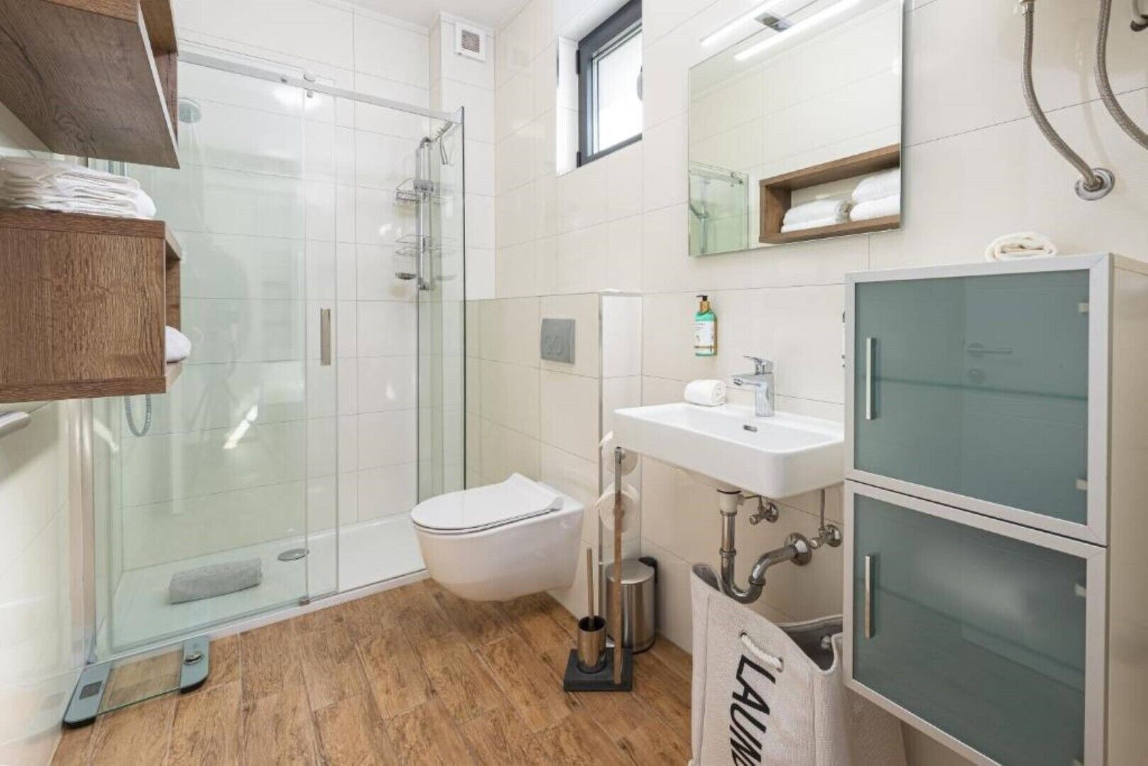 A modern bathroom with a glass shower enclosure, a toilet, a sink with a mirror, and a wooden shelf.