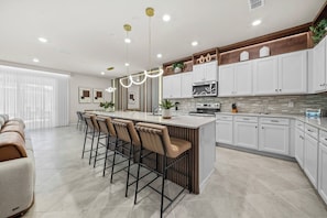 A modern kitchen with a central island, bar stools, white cabinetry, and stainless steel appliances. - 10BR house at Windsor Cay Resort (Clermont)