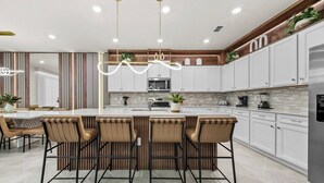 A modern kitchen with a central island, bar stools, and a dining area with a table and chairs. - 10BR house at Windsor Cay Resort (Clermont)