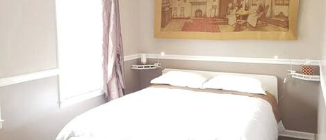 A bedroom with a large bed, a window with curtains, a wall-mounted painting, and a radiator.