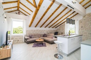 A modern loft with exposed wooden beams, a stone wall, a sofa, a coffee table, and a kitchen area. - Three Bedrom Apartment (Slano)