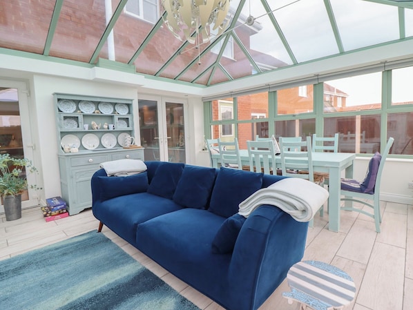 A conservatory with a blue sofa, dining table, and chairs, and a display of plates and cups.