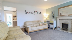 A living room with a beige sofa, a patterned armchair, a small side table, a bookshelf, and a fireplace. - The Beach House (Prestatyn)