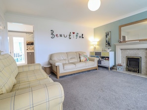 A living room with a beige sofa, a patterned armchair, a small side table, a bookshelf, and a fireplace. - The Beach House (Prestatyn)