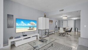 Ein modernes Wohnzimmer mit einem großen Flachbildfernseher, einer weißen Unterhaltungsanlage, einem Glastisch und einem Essbereich mit einer Pendelleuchte.