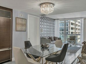 A modern living room with a glass dining table, a chandelier, and a view of a building through large windows. - Icon Brickell - 42nd Fl - Oceanview - Spa Access (Miami)