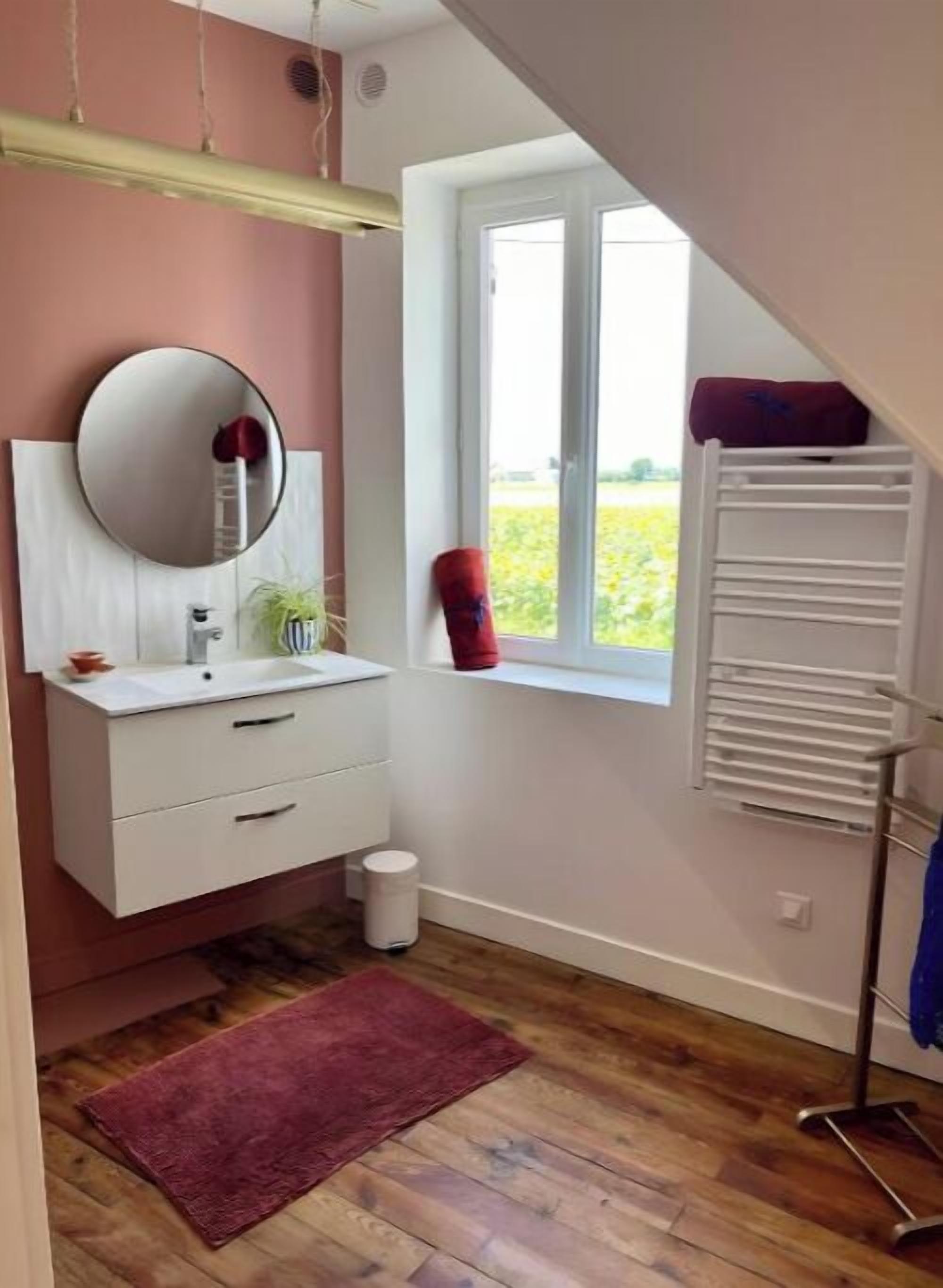 A bathroom with a white vanity, a round mirror, a window with a view of greenery, a red towel, and a white radiator.