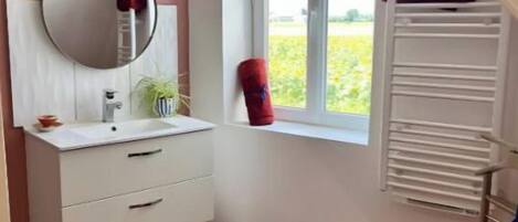 A bathroom with a white vanity, a round mirror, a window with a view of greenery, a red towel, and a white radiator.
