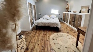 A bedroom with a sloped ceiling, a bed with patterned bedding, a wooden floor, a round woven rug, and a rustic wooden cabinet. - Les Hirondelles du Marais (Riom)