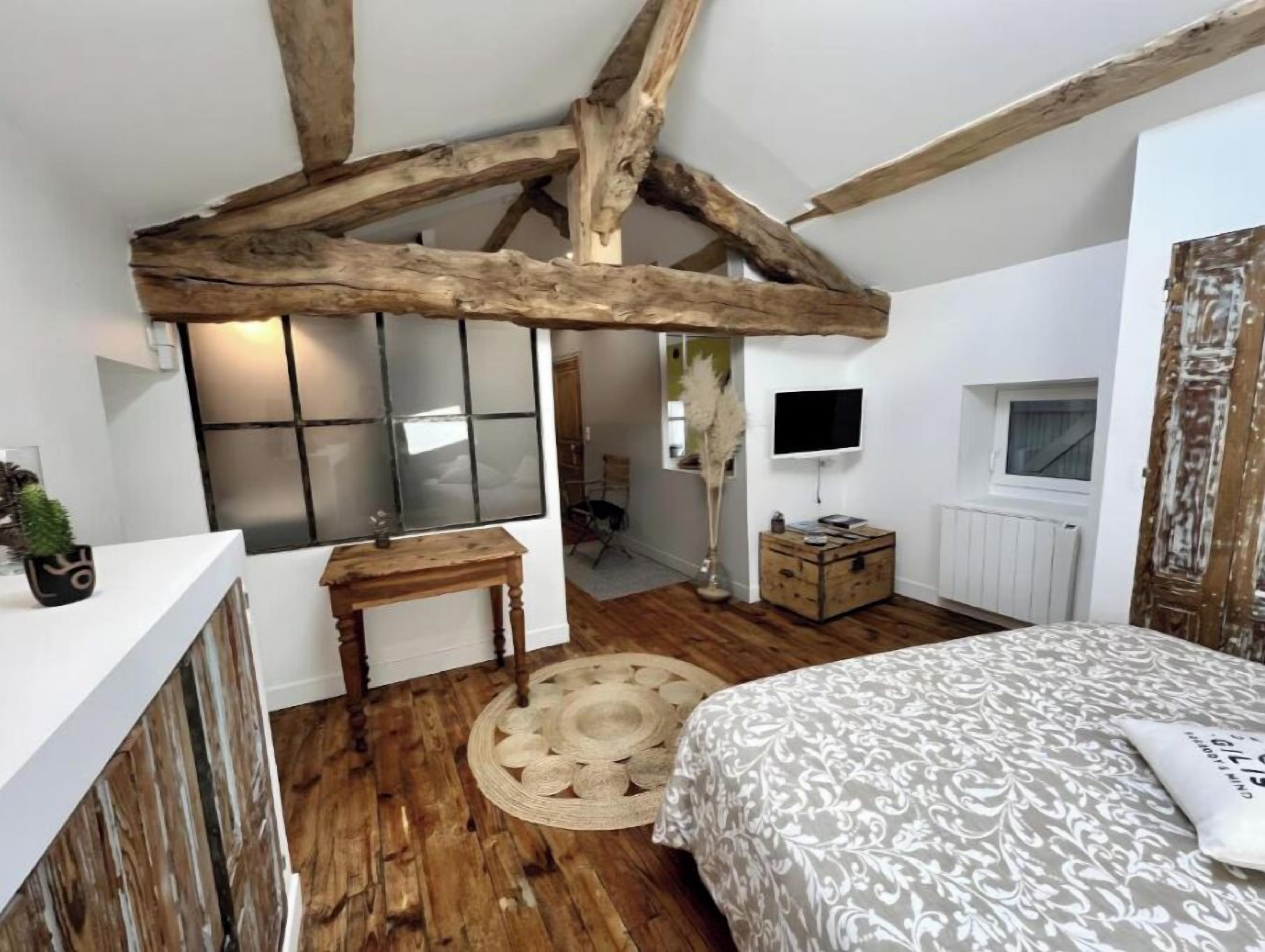 Un dormitorio con vigas de madera a la vista, una cama con colcha estampada, una pequeña mesa de madera, un espejo y un televisor en la pared.
