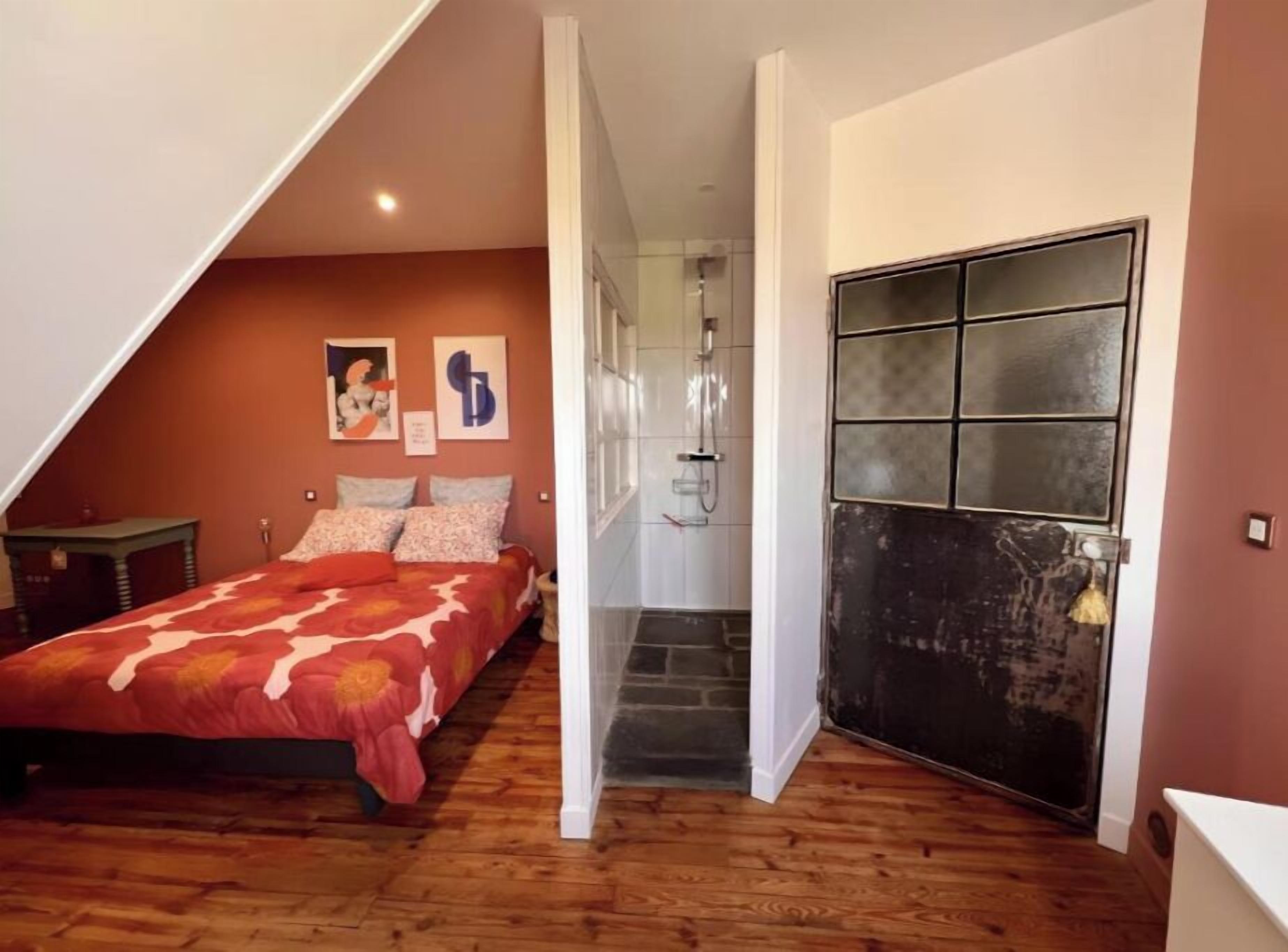 Une chambre avec un plafond en pente, un lit avec une literie à motifs floraux, une petite table et un espace douche.