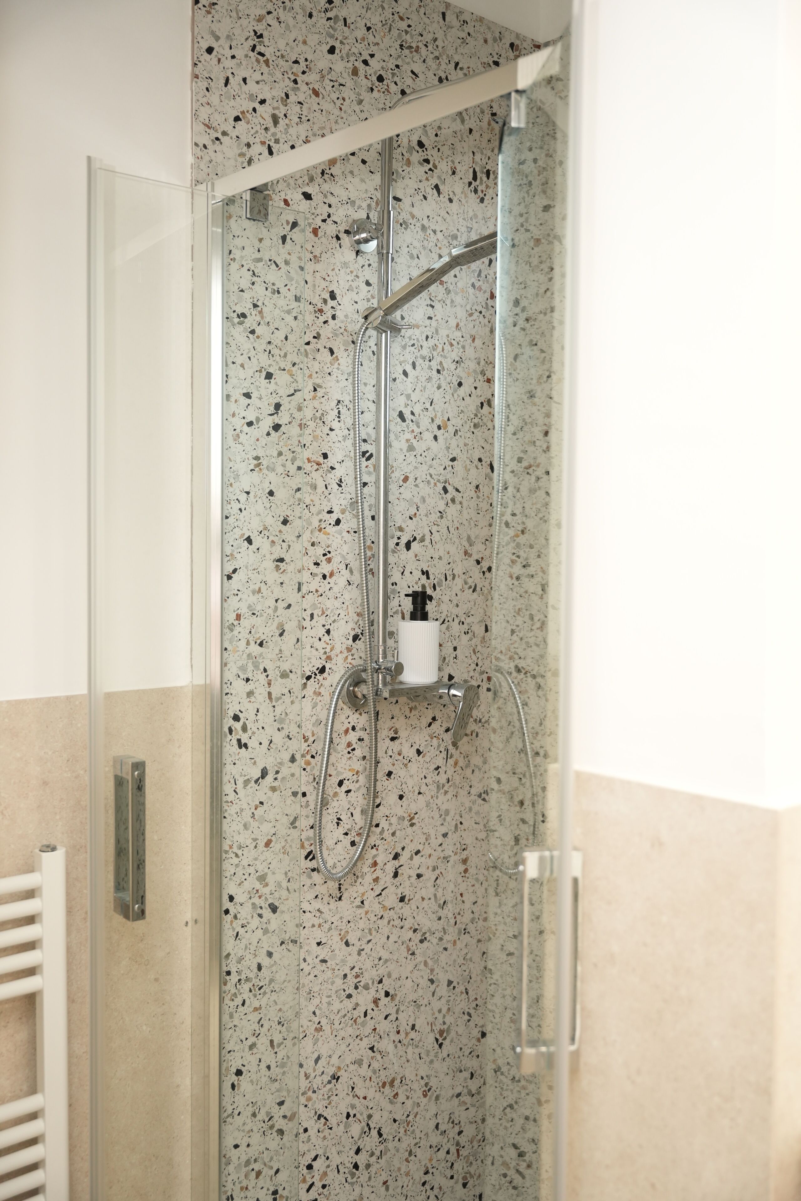 Une douche fermée par des parois de verre, avec un mur recouvert de carreaux à motifs, une douchette à main et un distributeur de savon.