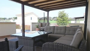 Een overdekt terras met rieten meubels, een tafel met glazen blad en een hangstoel.