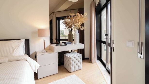 A bedroom with a bed, bedside table, lamp, dresser, vanity, and a large window.