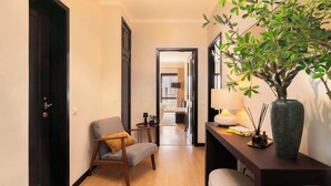 A hallway with a wooden floor, a chair with a cushion, a desk with a lamp, a vase with a plant, and a door.