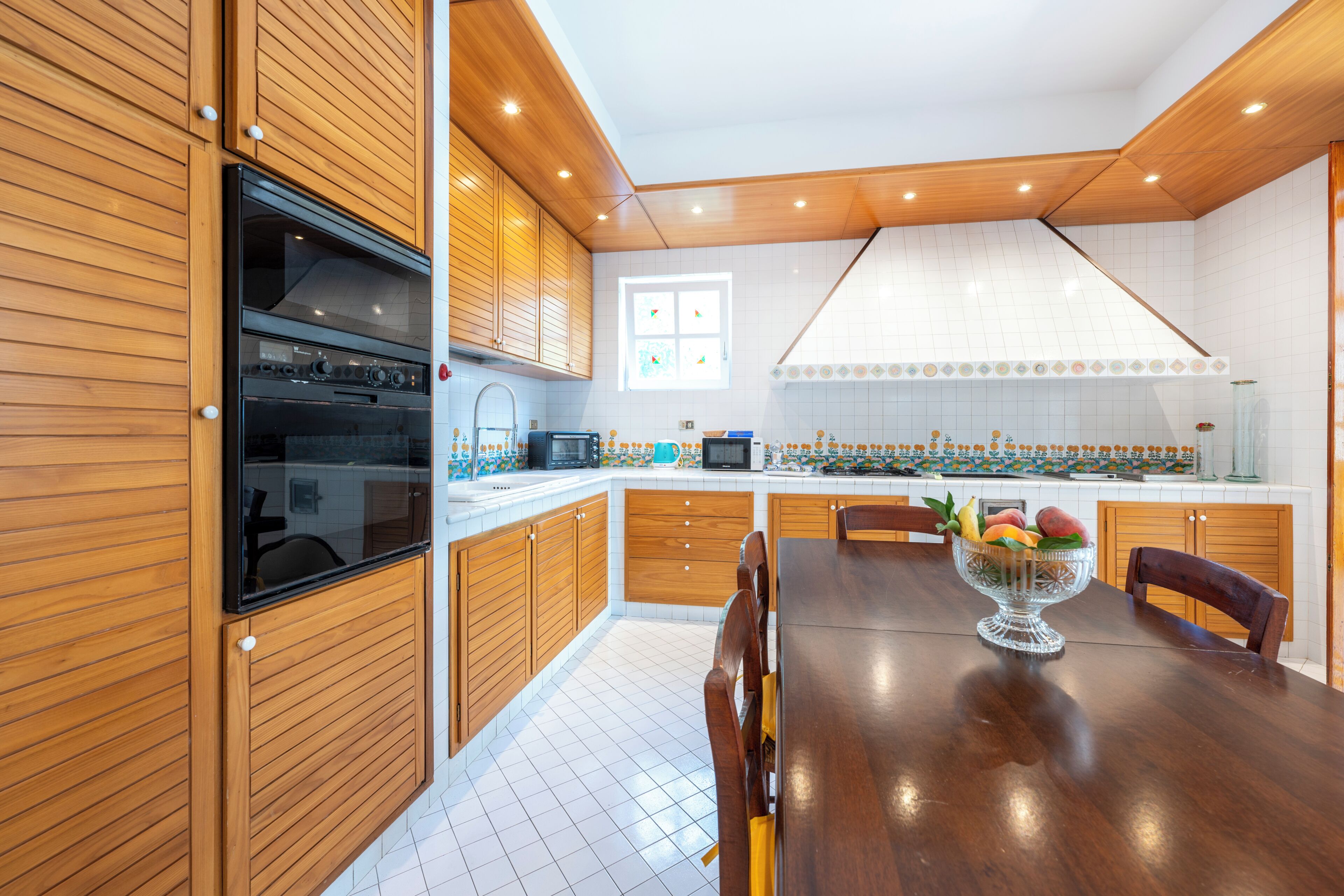 A modern kitchen with wooden cabinets, a built-in oven, a dining table with chairs, and a fruit bowl.
